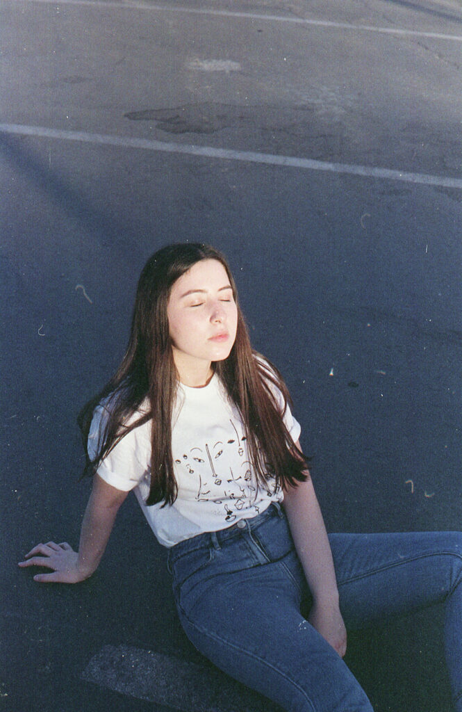 Film Photo of Gabriela Inzunza a 21-year-old from Los Angeles CA sitting on a parking lot during the golden hour. She wears a white t-shirt with a graphic design and blue jeans.